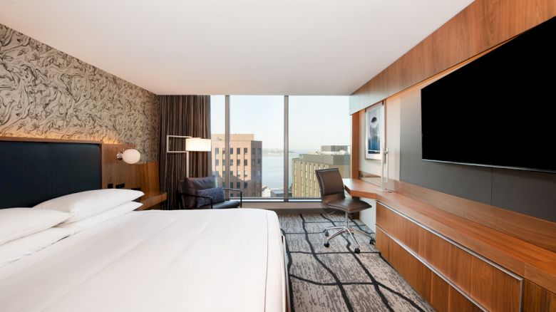 A modern hotel room with a large bed, wall-mounted TV, desk, chair, and a window with a cityscape view in the background.