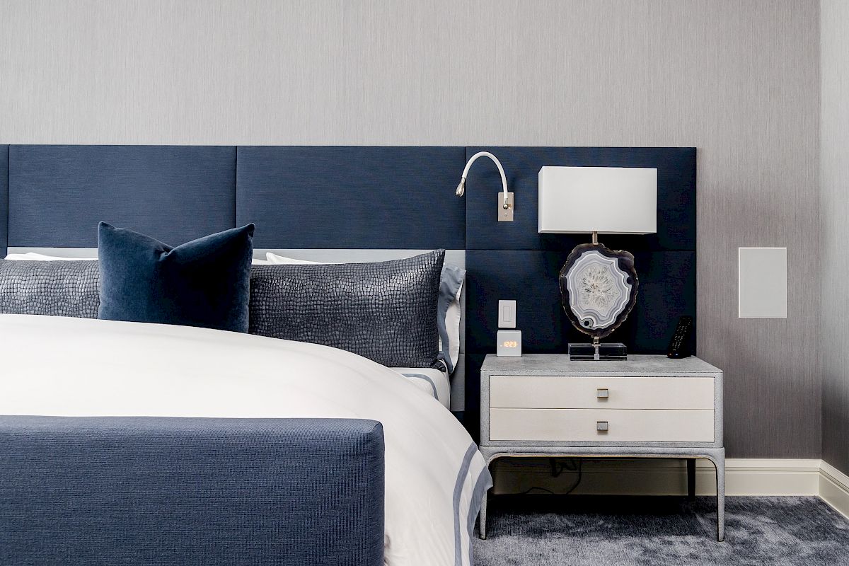This image shows a modern bedroom with a navy blue bed, white linens, a side table, and a lamp. The room features a grey wall and carpet.