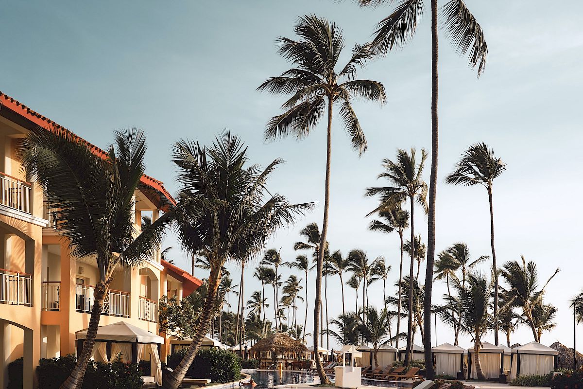 A serene poolside scene with lounge chairs, tall palm trees, and a building in the background under a clear sky, exuding tropical vacation vibes.