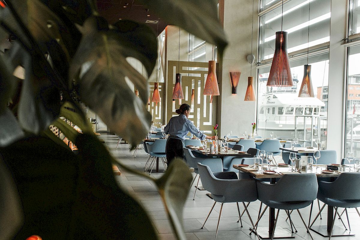 A restaurant with modern decor, featuring blue chairs, wooden tables, and large window views. A person is setting up a table.