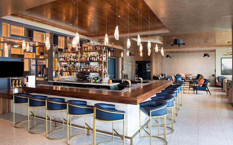 The image shows a modern bar with blue cushioned stools, a well-stocked liquor shelf, hanging lights, and a seating area in the background.