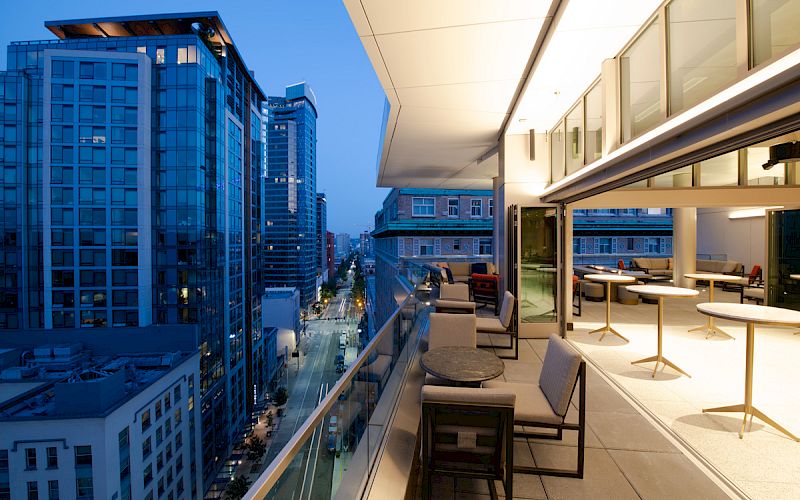 A modern cityscape at dusk featuring tall buildings and a spacious balcony with tables and chairs overlooking the street below.