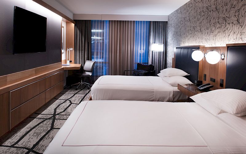 A modern hotel room with two double beds, a flat-screen TV, a desk, and modern decor featuring a patterned wall and curtains.