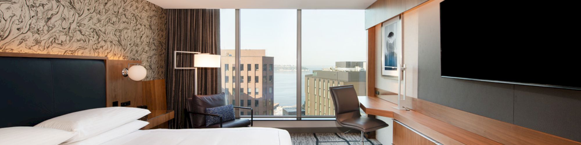 A modern hotel room with a large bed, wall-mounted TV, desk, and chair. There's a floor-to-ceiling window offering a view of buildings and water.